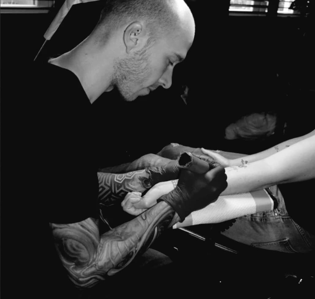 Bald tattoo artist with sleeve tattoos carefully inks a client's forearm in black and white.